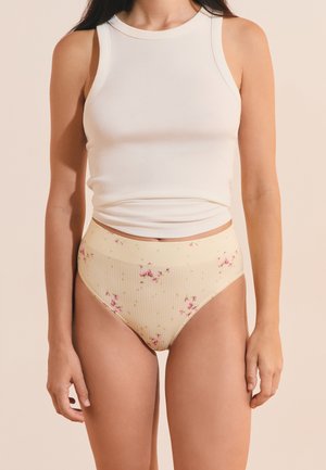 Donna che indossa un top corto bianco senza maniche a coste e mutandine beige a vita alta con una piccola stampa floreale rosa.