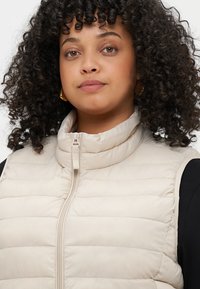 Smėlinis pūkinis liemenė su aukštu apykakle, turi priekinį užtrauktuką, siuvinėtą dizainą ir lygią tekstūrą. Dėvima ant juodos palaidinės.