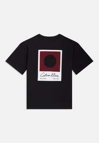 Černé bavlněné tričko s grafickým designem zahrnujícím čtverec s červenými pruhy a centrálním kruhem, pod nímž je nápis "Calvin Klein New York".