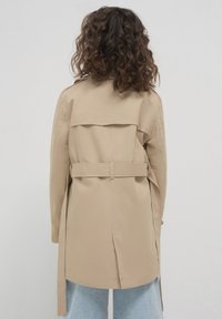 Beige Trenchcoat mit gebundenem Gürtel, Rückenpasse und Belüftung. Hat einen lockeren Schnitt und lange Ärmel. Wird über hellblauen Denim getragen.