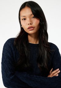 Granatowy sweter warkoczowy z teksturowanym wzorem, z grubymi prążkowanymi mankietami i okrągłym dekoltem. Noszony przez modelkę z długimi ciemnymi włosami i delikatnymi kolczykami.