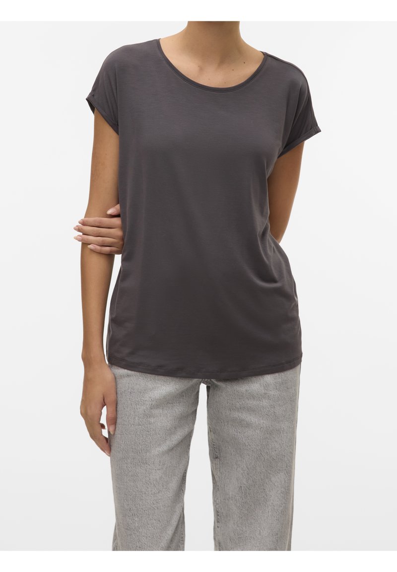 Femme portant un t-shirt gris foncé à manches courtes et un jean gris clair, debout avec un bras croisé sur sa taille.