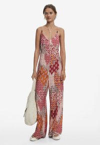 Mono fluido con un patrón de patchwork que presenta rojos, rosas y naranjas. Tirantes finos, escote en V, piernas anchas y acompañado de un bolso tejido.