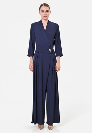 Donna che indossa una tuta blu navy a maniche lunghe con pantaloni ampi, davanti incrociato e cintura con fibbia dorata, abbinata a scarpe a punta color nude.