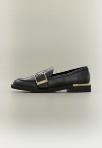 Mocasines de cuero negro con una gran hebilla dorada y un tacón bajo en bloque con un detalle dorado. El diseño es elegante y aerodinámico.