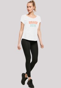 Weißes T-Shirt mit "BEACH BUM" Grafik in Orange und Blau, kombiniert mit schwarzen Leggings und schwarzen Sneakers. Lässiges, entspanntes Design.