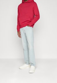 Sudadera roja con capucha y bolsillo frontal, con un pequeño logo, combinada con vaqueros de pierna recta en azul claro y zapatillas blancas.