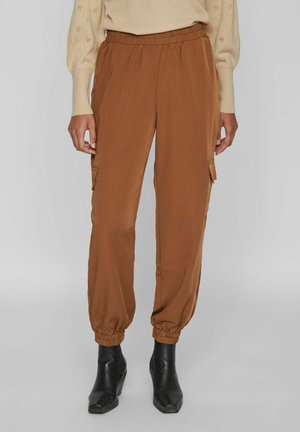 Braune Cargo-Hose mit elastischem Bund und geschnürten Bündchen, kombiniert mit schwarzen, spitz zulaufenden Stiefeletten und einem beigen Pullover.
