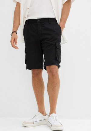 Shorts cargo noirs avec plusieurs poches latérales, fabriqués en tissu durable, portés avec un t-shirt blanc et des baskets blanches. Coupe décontractée.