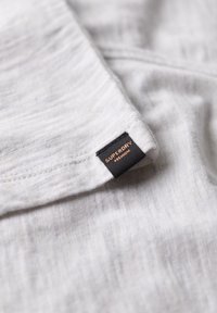 Lichtgrijze stof met een zachte textuur en een subtiel heatherpatroon. Voorzien van een zwart label met "SUPERDRY" in gouden letters.
