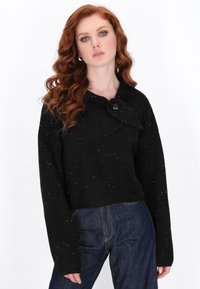 Zwart cropped sweater met een strikhalslijn en knoopdetails, met een gespikkelde textuur. Gecombineerd met donkerblauwe jeans.