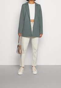 Šedé sako s výrazným střihem, nošené přes bílý crop top. Kombinováno s světle béžovými kalhotami a bílými teniskami. Drží malou hnědou kabelku.