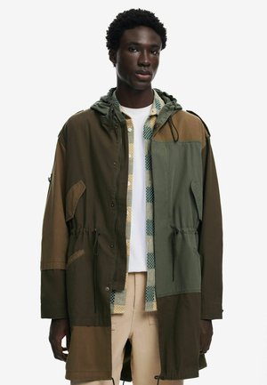 Oversized, two-tone jacket in olive green and brown tones, featuring a hood, drawstring waist, and side pockets. Textured fabric with contrasting sections.