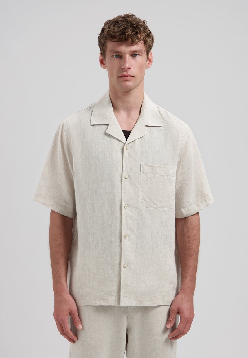 Jeune homme aux cheveux bouclés portant une chemise en lin beige à manches courtes avec boutons et un pantalon assorti, debout devant un fond uni.