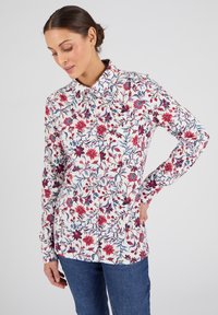 Donna con capelli scuri raccolti a chignon, che indossa una camicia bianca a maniche lunghe con motivi floreali rossi e blu e jeans blu, in posa su uno sfondo neutro.