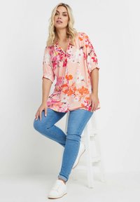 Blusa floreale rosa con fiori rossi e bianchi, dotata di colletto e maniche corte, abbinata a jeans skinny blu e sneakers bianche.