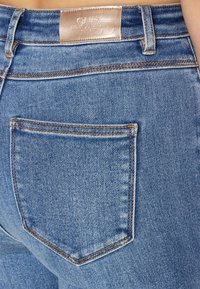 Jeans de denim azul claro con un ajuste de tiro medio, que presentan un único bolsillo trasero y una etiqueta de cuero en la cinturilla. Detalles de costura visibles.