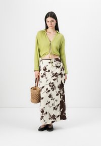 Chemise verte texturée à manches longues avec cordon de serrage, jupe maxi à motifs floraux en crème et marron, et un sac en osier. Chaussures plates noires.