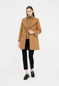 Cappotto cammello con collo a revers, doppiopetto con bottoni e tasche laterali, abbinato a un dolcevita marrone e pantaloni neri aderenti. Mocassini neri.