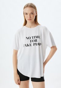 Hvid kortærmet t-shirt med rund halsudskæring, prydet med teksten "NO TIME FOR FAKE PEOPLE" i sort, tekstureret stof, parret med sorte shorts.