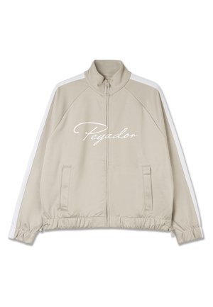 Beige zip-up jakke med hvite detaljer, har høy krage, elastiske mansjetter, snorjusterbar nederkant, og "Pegador"-logoen på forsiden.