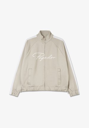 Biskvito spalvos zip-up striukė su baltais akcentais, turinti aukštą apykaklę, elastingus rankogalius, raišteliu reguliuojamą apačią ir "Pegador" logotipą priekyje.
