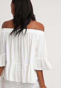 Bílý top bez ramínek s krátkými volánkovými rukávy a uvolněným střihem. Vyrobeno z lehkého materiálu s hladkou texturou.