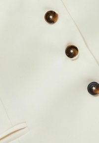 Tessuto color crema con una texture liscia, caratterizzato da bottoni circolari marroni come dettagli hardware sul davanti.