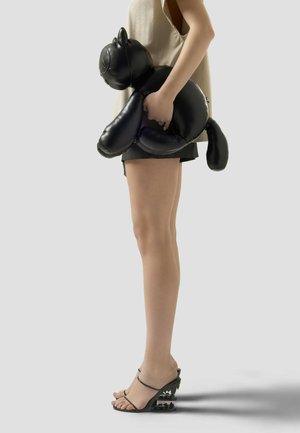 Persona con camiseta beige y pantalones cortos negros sosteniendo un bolso negro en forma de gato, usando sandalias negras con tiras y tacones gruesos esculpidos.