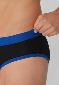 Schwarze und blaue Boxershorts mit einem kontrastierenden blauen elastischen Bund, glattem Stoff und passgenauer Passform, mit einer Hand, die den Bund greift.