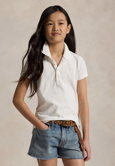 Jeune fille aux longs cheveux foncés porte un polo blanc, un short en jean et une ceinture tressée marron, debout avec une main dans la poche.