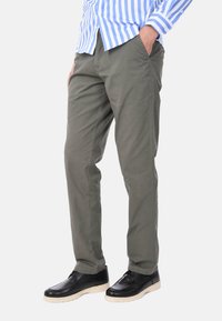 Pantaloni in cotone verde con design a gamba dritta, dotati di due tasche laterali, indossati con scarpe nere e una camicia a righe blu e bianche.