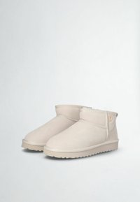 Stivali alla caviglia in suede beige con una texture morbida, punta rotonda, dettagli cuciti e un piccolo emblema logo sul lato, poggiati su una suola piatta.