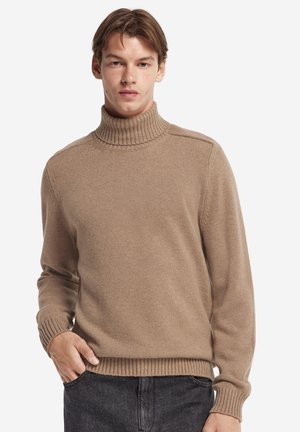 Ung mand iført en beige rullekravesweater og mørke bukser, stående med den ene hånd i lommen foran en ensfarvet baggrund.