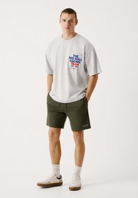 Graues, übergroßes T-Shirt mit blauer und roter Schrift "DAS RED BULL RACING TEAM EST. 2004", kombiniert mit grünen Shorts und weißen Sneakern.