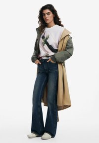 Cazadora puffer verde oliva superpuesta sobre un abrigo trench beige, camiseta gráfica blanca, jeans ajustados de mezclilla oscura y zapatillas blancas sin cordones.