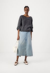 Grijze gebreide trui met pofmouwen, gecombineerd met een lichtblauwe denim maxi rok. Het model houdt een witte handtas vast en draagt beige schoenen.