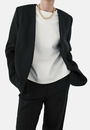 Personne portant un costume noir avec un haut blanc et un collier épais de perles dorées, mains dans les poches, sur un fond clair uni.