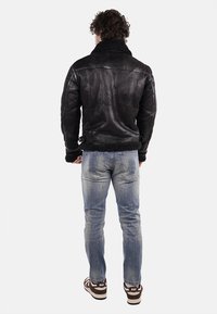 Giubbotto di pelle nera con finitura texturizzata e colletto in pelliccia. Indossato con jeans chiari strappati e sneakers marroni. Dettagli di cucitura visibili.