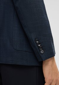 Close-up van een donkerblauwe gestructureerde blazer mouw met vier zwarte knopen, gedragen met zwarte broeken en toont een deel van een arm van een persoon.
