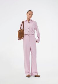 Φόρεμα σε ανοιχτό ροζ χρώμα με κουμπιά και δύο τσέπες στο στήθος, συνδυασμένο με ανοιχτό ροζ παντελόνι σε χαλαρή γραμμή. Η εμφάνιση ολοκληρώνεται με καφέ τσάντα ώμου και καφέ παπούτσια.