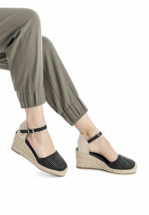 Zoccoli espadrille intrecciati neri con tacco testurizzato beige, dotati di cinturino alla caviglia e suola piatta per maggiore supporto.