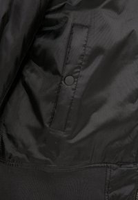 Veste noire en tissu lisse et léger. Comprend un bouton pression sur une poche latérale, des poignets côtelés et une finition texturée.