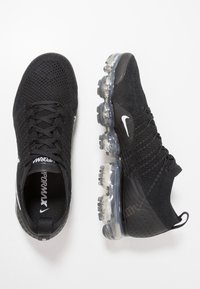 Chaussures de sport noires avec une tige en maille respirante, surface texturée et amorti d'air visible dans la semelle. Présente un logo blanc et des lacets.