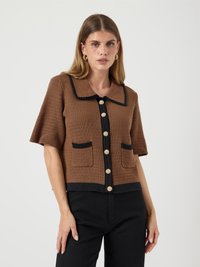 Mulher a usar um cardigan tricotado de mangas curtas castanho com detalhes pretos, botões dourados, bolsos frontais e calças pretas, contra um fundo branco liso.
