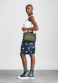 Alyvuogių žalios spalvos maišelis su įspaustu tekstu "THE TOTE BAG" auksinės spalvos. Dėvimas su džinsine sijonine, puošta baltomis gėlėmis, ir mėlynomis sportinėmis aukštakulnėmis.