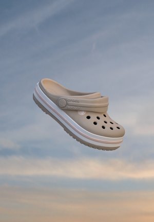 Sabot Crocs beige avec des trous d’aération et une semelle blanche et rose, flottant sur fond de ciel nuageux.