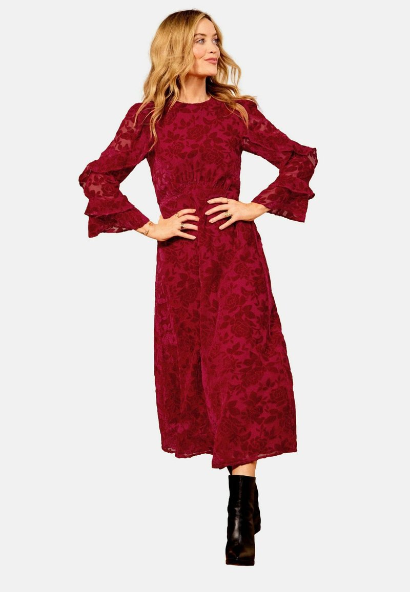 Burgunderfarbendes Spitzenkleid mit langen, rüschigen Ärmeln und tailliertem Schnitt. Kombiniert mit schwarzen Stiefeletten. Midi-Länge mit fließender Textur.