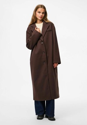 Giovane donna che indossa un lungo cappotto marrone abbottonato, un maglione crema, jeans larghi blu e stivali neri, in piedi davanti a uno sfondo bianco semplice.