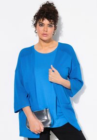 Blå cardigan med avslappnad passform, med en texturerad stickning på axlarna och fickorna. Bärs över en matchande blå topp och kombineras med en silverclutch.